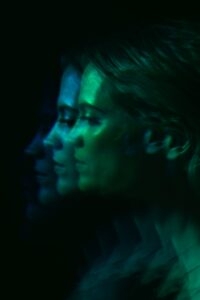 The same woman's profile repeated three times in overlapping layers, lit in deep green and teal against a black background.